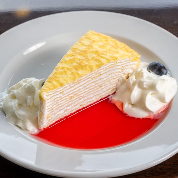 Slice of crepe cake with whipped cream, a blueberry, and red sauce on a white plate.