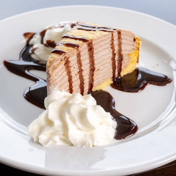 A slice of chocolate layered cake with chocolate drizzle and whipped cream on a white plate.
