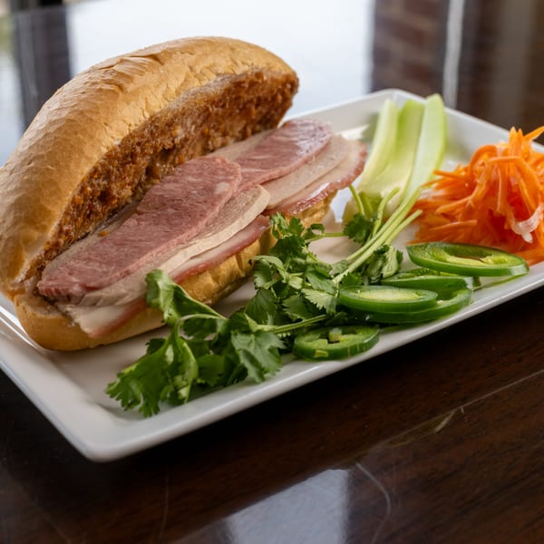 A Vietnamese banh mi sandwich with sliced meats, cilantro, jalapenos, and pickled carrots on a white plate.