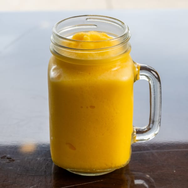 A vibrant yellow mango smoothie in a glass mason jar mug on a dark wooden table.