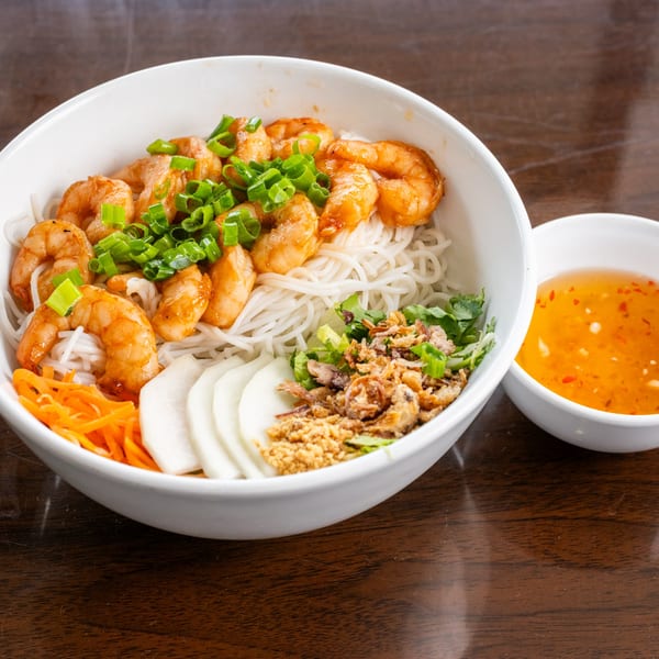 A bowl of Vietnamese vermicelli noodles with shrimp, scallions, and shredded vegetables, with a side of sauce.